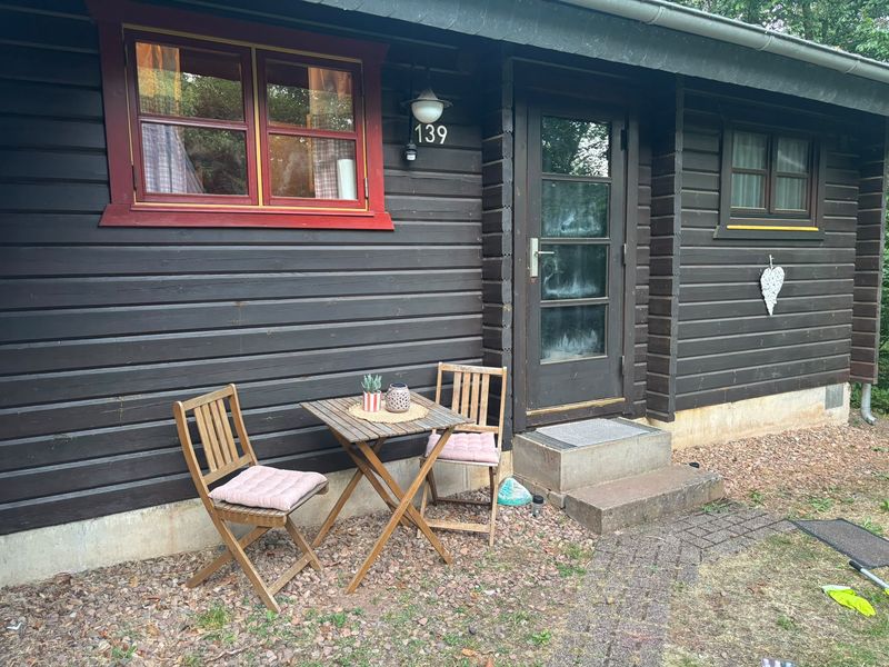 Vorderseite des dunklen Holzhauses mit Hausnummer 139, roten Fensterrahmen und Bistrotisch