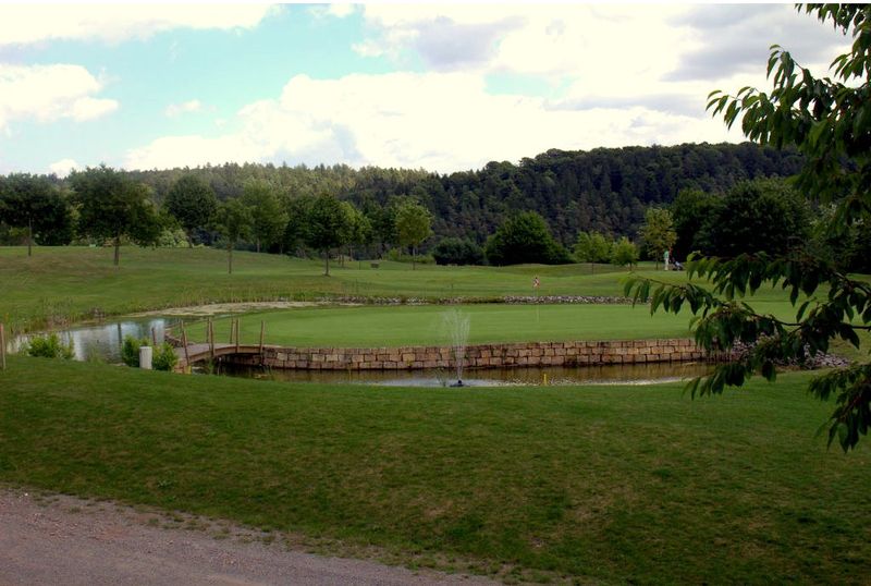 Golfplatz mit Green, Teich und kleiner Holzbrücke vor bewaldeten Hügeln