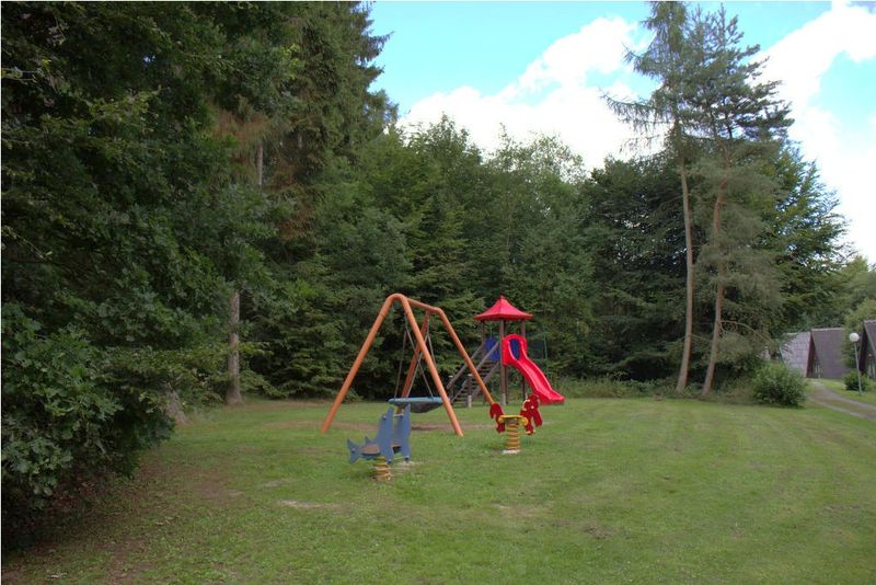 Spielplatz mit Schaukel, roter Rutsche und Federwippen auf einer Wiese am Waldrand